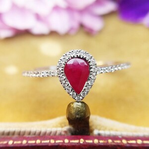May include: A silver ring with a pear-cut, deep red ruby gemstone, encircled by a halo of small, clear diamonds. The ring's band is also set with tiny diamonds, creating a sparkling effect. The ring is set against a soft, yellow background.