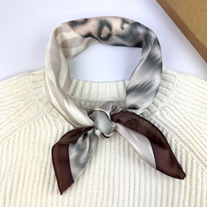 May include: A square silk scarf featuring a mix of patterns including leopard print, abstract grey brushstrokes, and solid dark brown borders. The scarf is tied in a knot and rests against a cream-coloured ribbed knit sweater with a high neckline.
