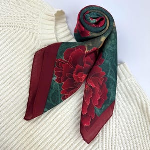 May include: A square silk scarf featuring a deep red floral pattern on a dark green background with a solid burgundy border. The scarf is partially rolled and displayed against a cream-coloured ribbed knit sweater, showcasing the fabric texture.