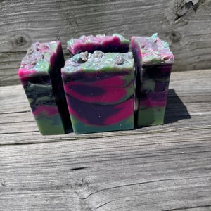 Könnte beinhalten: Vier handgemachte Seifenstücke mit einem geschichteten Design. Die Seife weist Wirbel aus Magenta, Grün und Schwarz auf. Die Oberseite jeder Seife hat ein strukturiertes, blütenartiges Design. Die Seifen werden auf einer verwitterten Holzoberfläche präsentiert.