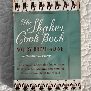 May include: A vintage cookbook titled "The Shaker Cook Book" by Caroline B. Piercy. The cover is teal and white with illustrations of Shaker figures. The book's title and subtitle, "NOT BY BREAD ALONE," are prominently displayed. The cover also includes text about Shaker recipes.