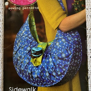 May include: A sewing pattern for a blue and green fabric satchel bag with a floral embellishment. The pattern is called "Sidewalk Satchel" and is from the brand "Anna Maria Sewing Patterns".