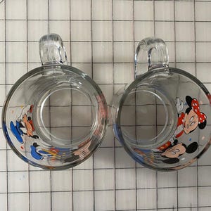 Puede incluir: Dos tazas de vidrio transparente con cuerpos redondeados y asas transparentes. Cada taza presenta un dise&ntilde;o de dibujos animados colorido de Mickey y Minnie Mouse. Las tazas est&aacute;n sobre un fondo de cuadr&iacute;cula blanca.