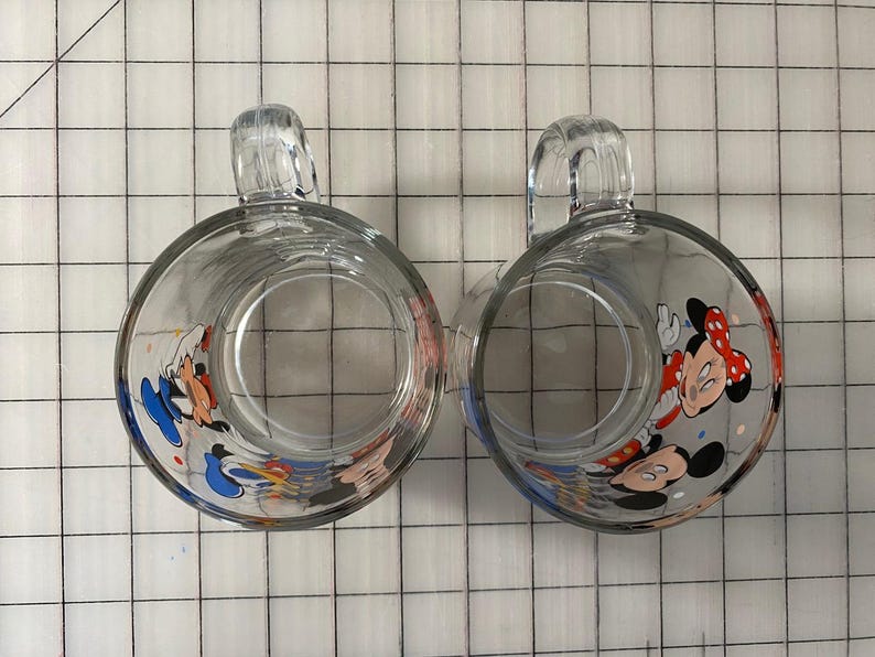 Puede incluir: Dos tazas de vidrio transparente con cuerpos redondeados y asas transparentes. Cada taza presenta un dise&ntilde;o de dibujos animados colorido de Mickey y Minnie Mouse. Las tazas est&aacute;n sobre un fondo de cuadr&iacute;cula blanca.