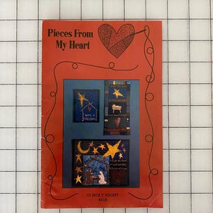 May include: A red book titled "Pieces From My Heart" with a heart design and three framed images of stars and religious scenes. The text "O' Holy Night" and "#115" are at the bottom. The book is on a white grid surface.