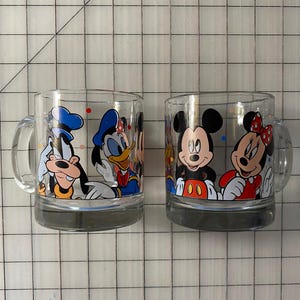 Puede incluir: Dos tazas de vidrio transparente con personajes de Disney. Una taza muestra a Goofy y el Pato Donald, mientras que la otra presenta a Mickey y Minnie Mouse. Cada taza tiene un asa y una base gris, ideal para los fan&aacute;ticos de los dibujos animados cl&aacute;sicos.