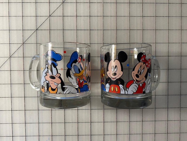 Puede incluir: Dos tazas de vidrio transparente con personajes de Disney. Una taza muestra a Goofy y el Pato Donald, mientras que la otra presenta a Mickey y Minnie Mouse. Cada taza tiene un asa y una base gris, ideal para los fan&aacute;ticos de los dibujos animados cl&aacute;sicos.