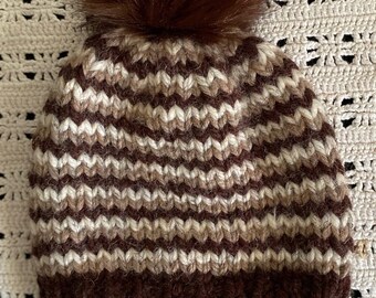 Hand-knit striped toddler size beanie hat in shades of brown topped with a brown faux pompom