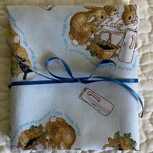 May include: A light blue fabric item tied with a dark blue ribbon. The fabric features illustrations of rabbits, baskets, and milk jugs, with text that reads "Flopsy, Mopsy, and Cotton-tail had bread and milk and blackberries."