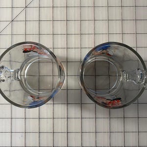 Puede incluir: Dos tazas de vidrio transparente con asas, cada una con un dise&ntilde;o de personaje de dibujos animados. Las tazas transparentes permiten ver el interior. Las asas son robustas y f&aacute;ciles de agarrar. Las tazas est&aacute;n colocadas sobre una superficie cuadriculada.