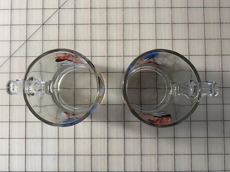Puede incluir: Dos tazas de vidrio transparente con asas, cada una con un dise&ntilde;o de personaje de dibujos animados. Las tazas transparentes permiten ver el interior. Las asas son robustas y f&aacute;ciles de agarrar. Las tazas est&aacute;n colocadas sobre una superficie cuadriculada.