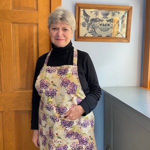 May include: A floral apron with a beige background and purple flower clusters. The apron has a pocket and is worn over a black turtleneck. A framed picture with a bird design hangs on the wall.