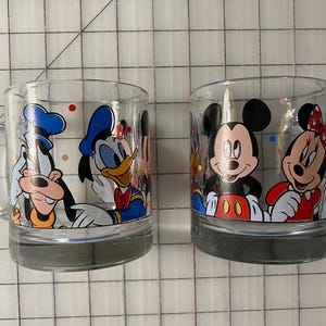 Puede incluir: Dos tazas de vidrio transparente con personajes de Disney. Una taza muestra a Goofy y el Pato Donald, mientras que la otra muestra a Mickey y Minnie Mouse. Cada taza tiene un asa y una base gris, perfectas para bebidas.