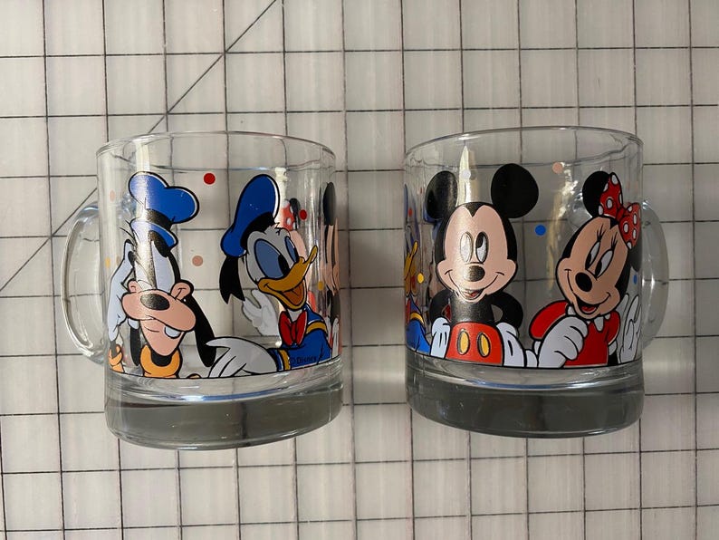 Puede incluir: Dos tazas de vidrio transparente con personajes de Disney. Una taza muestra a Goofy y el Pato Donald, mientras que la otra muestra a Mickey y Minnie Mouse. Cada taza tiene un asa y una base gris, perfectas para bebidas.