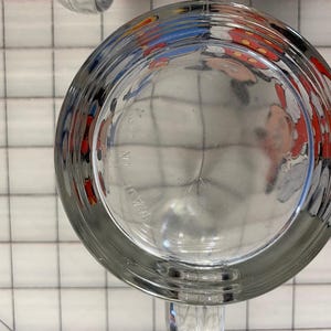 Puede incluir: Vaso de vidrio transparente con un borde ancho y acanalado. La parte inferior del vaso refleja una imagen colorida de un personaje de dibujos animados. Las palabras "MADE IN" son visibles en la parte inferior del vaso.