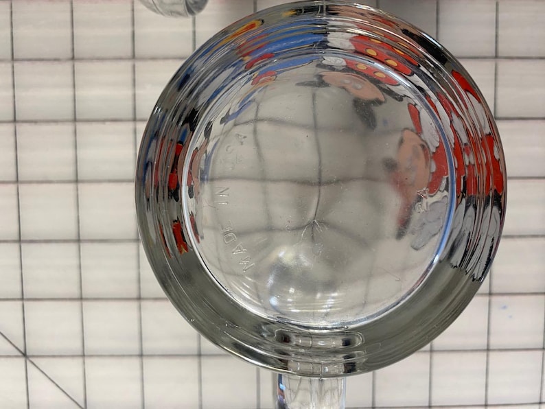 Puede incluir: Vaso de vidrio transparente con un borde ancho y acanalado. La parte inferior del vaso refleja una imagen colorida de un personaje de dibujos animados. Las palabras "MADE IN" son visibles en la parte inferior del vaso.
