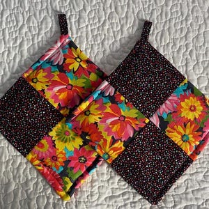 May include: Two square potholders with a patchwork design. One side features a black background with small, colorful polka dots. The other side has a vibrant floral pattern in shades of pink, yellow, and orange. Each potholder has a small fabric loop.