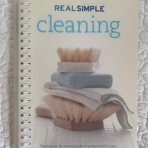 May include: A spiral-bound book titled "REAL SIMPLE cleaning" with blue lettering. The cover features a stack of cleaning brushes, cloths, and a sponge. The tagline reads, "Your room-by-room guide to a beautiful house."