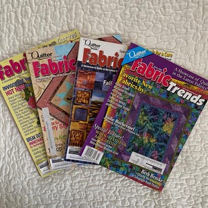 May include: Four colorful quilting magazines are fanned out on a white quilted surface. The magazines have titles like "Fabric Trends" and "Quilter's Fabric." The covers feature quilt patterns and articles about quilting.