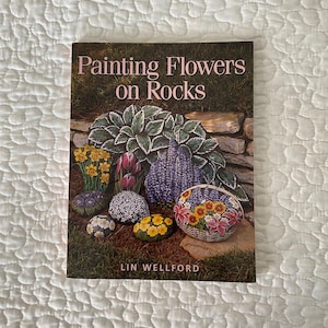 May include: A book titled "Painting Flowers on Rocks" featuring painted rocks with floral designs. The cover displays various painted rocks with flowers, including daffodils, tulips, and daisies. The author's name, Lin Wellford, is also visible.