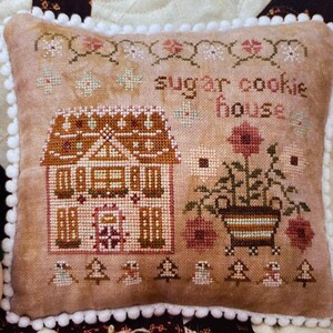 May include: A brown and tan embroidered pillow with a gingerbread house and the text "sugar cookie house".