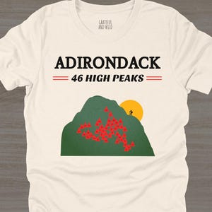 May include: Cream-colored t-shirt with the text "ADIRONDACK 46 HIGH PEAKS" in black. The graphic features a green mountain with red flowers, a yellow sun, and a small figure. The shirt is made of a soft material.