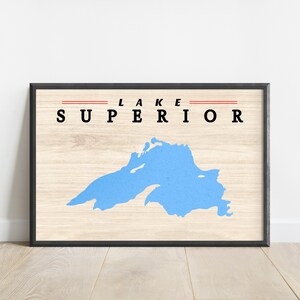 May include: Framed print featuring a light blue map of Lake Superior on a wood-grain background. The words "LAKE SUPERIOR" are printed in black, with a red line above the word "SUPERIOR". The frame is black.