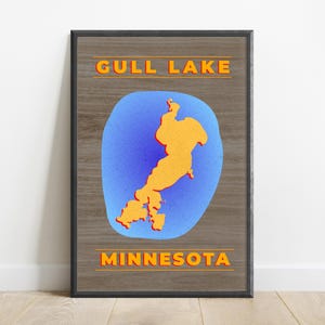 Puede incluir: Póster enmarcado con un mapa del lago Gull, Minnesota. El mapa es naranja sobre un fondo azul, con las palabras "GULL LAKE" y "MINNESOTA" en texto naranja. El marco es negro y el fondo tiene un patrón de vetas de madera.