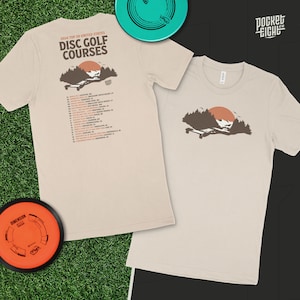 Può includere: Una maglietta beige con una grafica di un tramonto su una catena montuosa con alberi. Il testo "2023 TOP 20 UNITED STATES DISC GOLF COURSES" è stampato sul retro della maglietta.