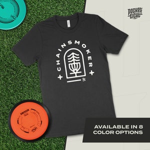 May include: Black t-shirt with a white graphic of a disc golf basket inside a tree with the text "Chainsmoker+" around it. The text "Pocket Eight Co." is in the top right corner of the image. The text "Available in 8 Color Options" is at the bottom of the image.