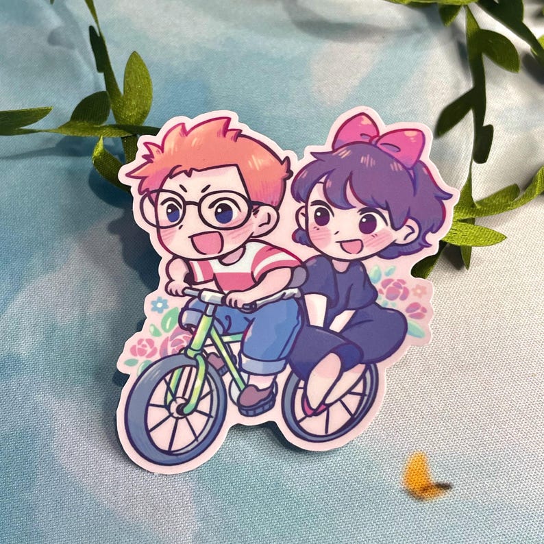 Op de afbeelding: Een sticker met twee cartoonfiguren die op een fiets rijden. Het personage vooraan draagt een rood-wit gestreept shirt en een blauwe spijkerbroek. Het personage achteraan draagt een zwarte jurk en een roze strik. De achtergrond is een roze-wit bloemenpatroon.