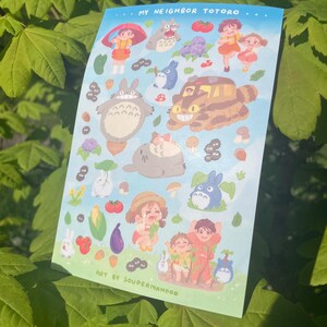 May include: A sticker sheet featuring illustrations of characters and elements from "My Neighbor Totoro." The sheet includes images of Totoro, Mei, Satsuki, and other iconic imagery from the animated film, set against a light blue background. Text includes "MY NEIGHBOR TOTORO" and "ART BY SOUPERMANDOO."