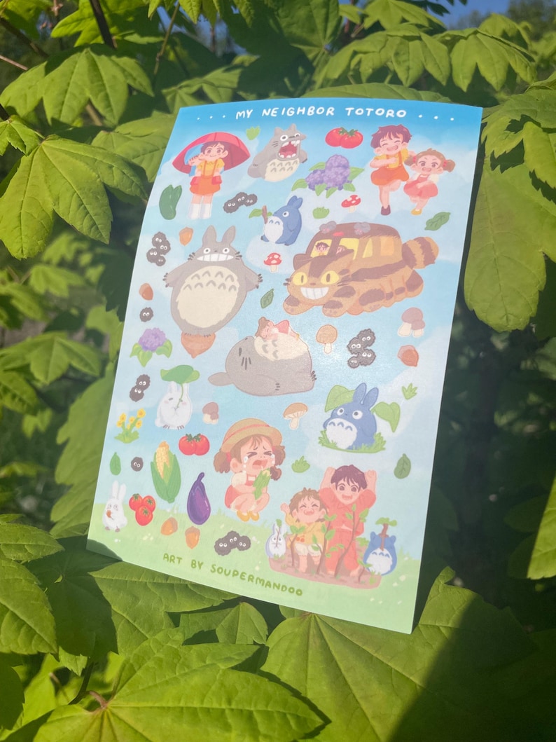 May include: A sticker sheet featuring illustrations of characters and elements from "My Neighbor Totoro." The sheet includes images of Totoro, Mei, Satsuki, and other iconic imagery from the animated film, set against a light blue background. Text includes "MY NEIGHBOR TOTORO" and "ART BY SOUPERMANDOO."