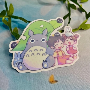 Op de afbeelding: Een sticker met een cartoon Totoro, twee kinderen en kleine zwarte roetgeesten. De Totoro zit op een groen blad en de kinderen lachen. De sticker is op een lichtblauwe achtergrond.