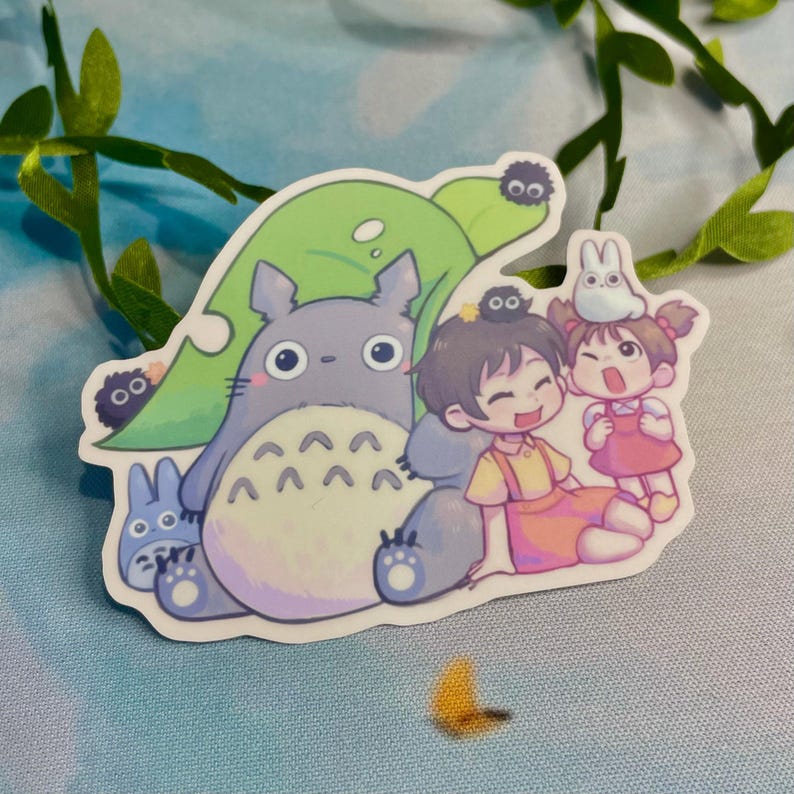 Op de afbeelding: Een sticker met een cartoon Totoro, twee kinderen en kleine zwarte roetgeesten. De Totoro zit op een groen blad en de kinderen lachen. De sticker is op een lichtblauwe achtergrond.