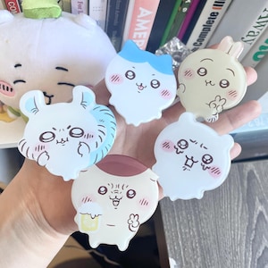 May include: A hand holds six cartoon character-shaped stickers. The stickers feature various designs, including a cat with blue accents, a bunny with blue accents, and other cute, round characters in white, beige, and brown. A plush toy is visible in the background.