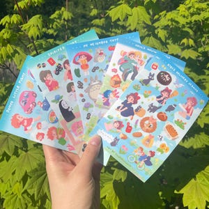 May include: A hand holding a stack of colorful sticker sheets featuring illustrations from animated films. The sheets have titles like "Ponyo Little Fishy," "Spirited Away," and "My Neighbor Totoro." The background is a lush green foliage.
