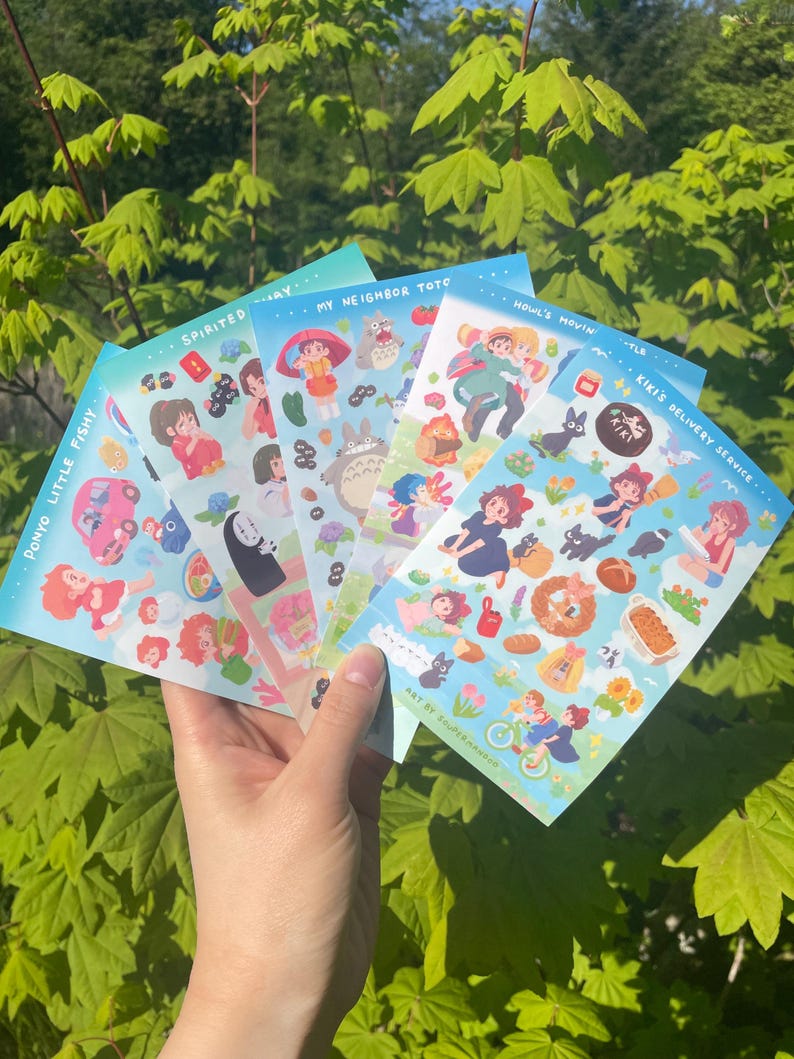May include: A hand holding a stack of colorful sticker sheets featuring illustrations from animated films. The sheets have titles like "Ponyo Little Fishy," "Spirited Away," and "My Neighbor Totoro." The background is a lush green foliage.