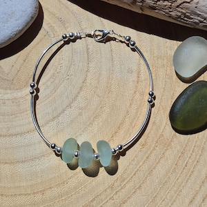 Puede incluir: Pulsera plateada con tres piezas de vidrio de mar verde pálido. La pulsera tiene un diseño minimalista con pequeñas cuentas plateadas y un cierre. El vidrio de mar tiene un aspecto liso y esmerilado.