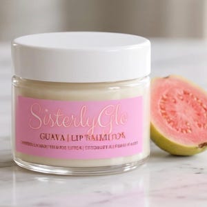 May include: A glass jar of Sisterly Glo Guava Lip Balm with a white lid and a pink label. A halved guava fruit with pink flesh sits beside the jar. The jar and fruit are on a marble surface.