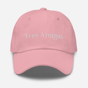 May include: A pink baseball cap with the text "Tres Amigas" embroidered in white on the front.