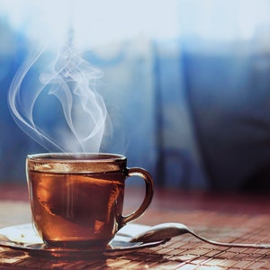 May include: A steaming cup of tea in a clear glass mug with a brown handle sits on a saucer on a woven placemat. The mug is filled with amber-colored tea.