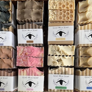 May include: A wooden crate filled with various handmade goat milk soaps. The soaps are in different colors and textures, with labels that read "Goat Milk Soap" and various scents like "Arctic Woods," "Espresso," and "Oat & Honey."