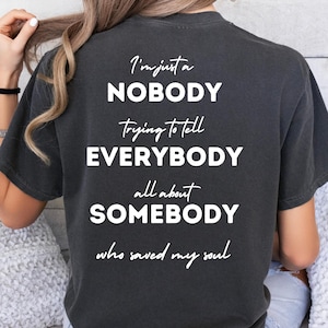 Könnte beinhalten: Dunkelgraues T-Shirt mit dem weißen Text "I'm just a NOBODY Trying to tell EVERYBODY all about SOMEBODY who saved my soul". Das T-Shirt hat einen lässigen Stil.
