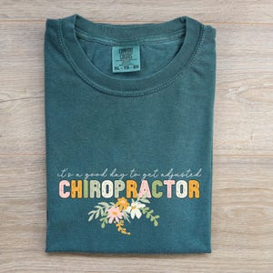 Peut inclure: T-shirt Comfort Colors teal avec le texte "it's a good day to get adjusted CHIROPRACTOR" en lettres colorées et un motif floral. Le t-shirt est de taille XL-TG-EG.
