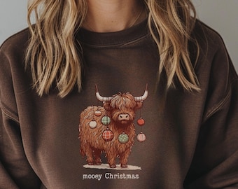 May include: Brown sweatshirt featuring a Highland cow illustration decorated with Christmas ornaments. The cow has long, shaggy brown fur and curved horns. The text "mooey Christmas" is printed below the cow in a white, sans-serif font.
