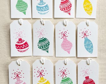May include: Twelve white gift tags with a red bow and a colorful ornament on each. The ornaments are in various colors, including red, green, blue, yellow, and pink.