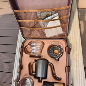Puede incluir: Un estuche marr&oacute;n con cremallera que contiene un juego para preparar caf&eacute;. El juego incluye una jarra de vidrio, un vertedor, una tetera de cuello de cisne, una taza de vidrio y un molinillo. El estuche tiene bolsillos de malla y correas marrones.