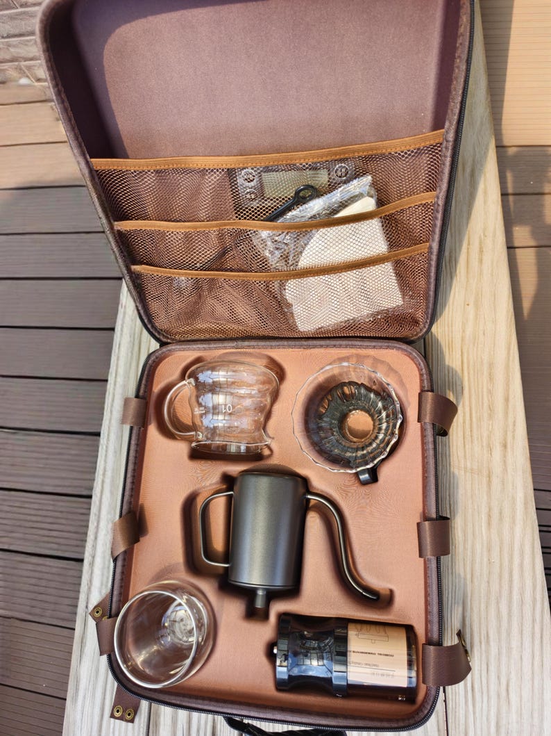 Puede incluir: Un estuche marr&oacute;n con cremallera que contiene un juego para preparar caf&eacute;. El juego incluye una jarra de vidrio, un vertedor, una tetera de cuello de cisne, una taza de vidrio y un molinillo. El estuche tiene bolsillos de malla y correas marrones.