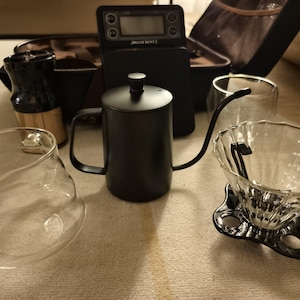 Puede incluir: Un conjunto de equipos para preparar caf&eacute;, que incluye una tetera de cuello de cisne negra, una jarra de vidrio, un gotero y una b&aacute;scula digital. Los art&iacute;culos est&aacute;n dispuestos sobre una superficie clara, lo que sugiere una preparaci&oacute;n de caf&eacute;.